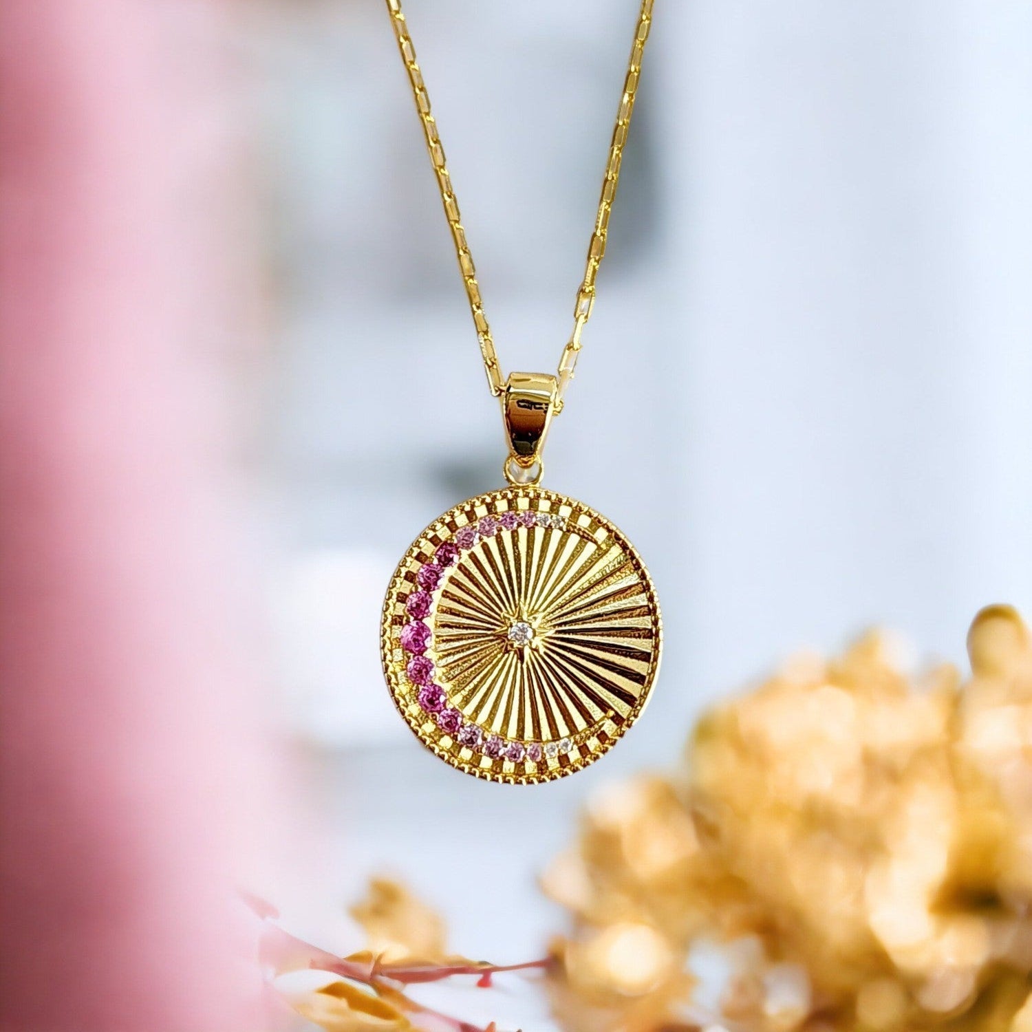 Saints and Saviors Gold necklace with a circular crescent moon and sun rays pendant featuring ombre pink crystals against a blurred natural background