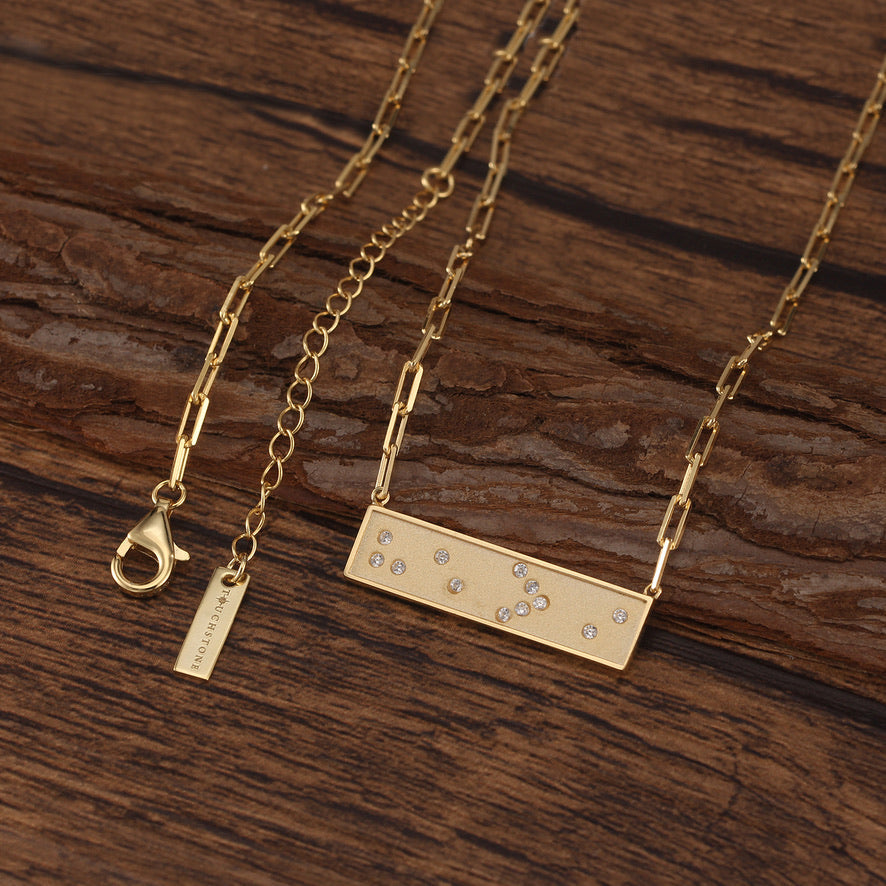 Gold Braille 'He > i' message in crystals on a rectangular pendant on gold link chain necklace on wooden surface