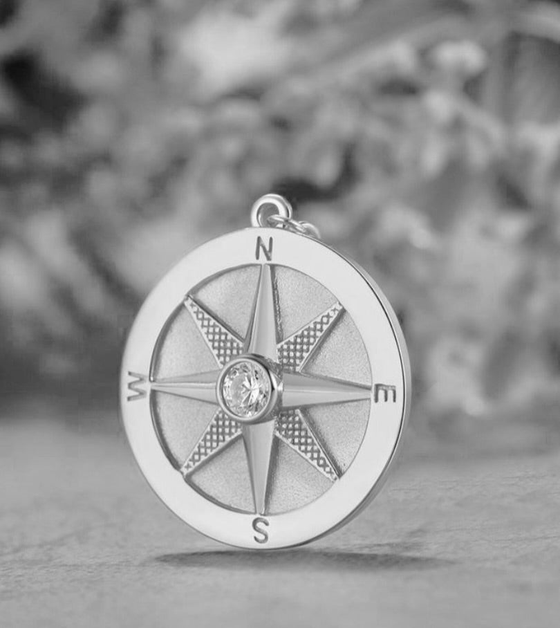 Silver compass pendant with a diamond in the center on a textured surface.