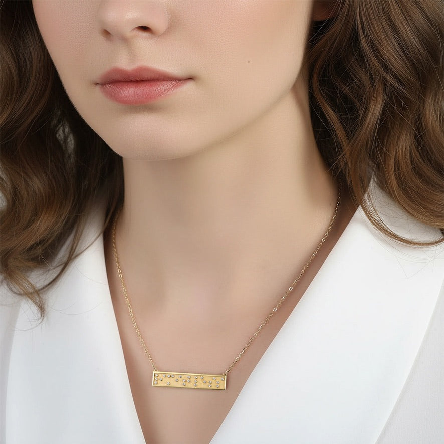 Gold necklace with a Limitless in Braille crystals rectangular pendant worn by a person in a white outfit.