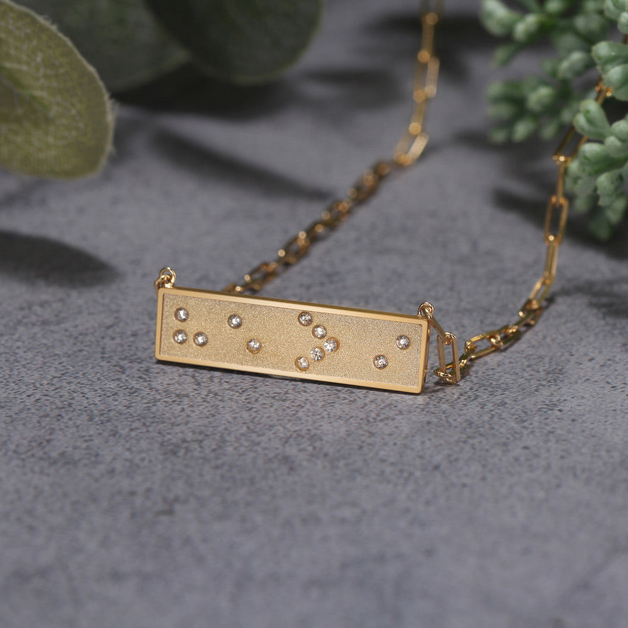 Gold necklace with a rectangular horizontal bar pendant featuring Braille He > i symbols on a gray surface with green leaves in the background.