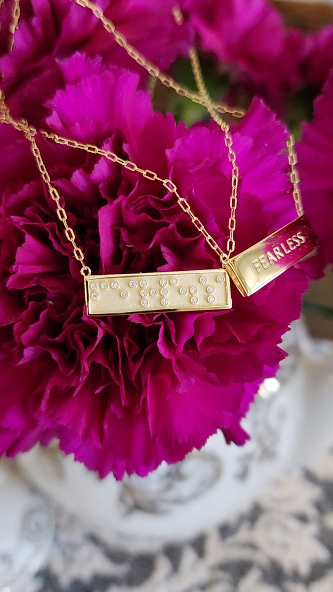 Gold mini bar necklace with 'FEARLESS' in Braille crystals and text on reverse side, on pink flowers