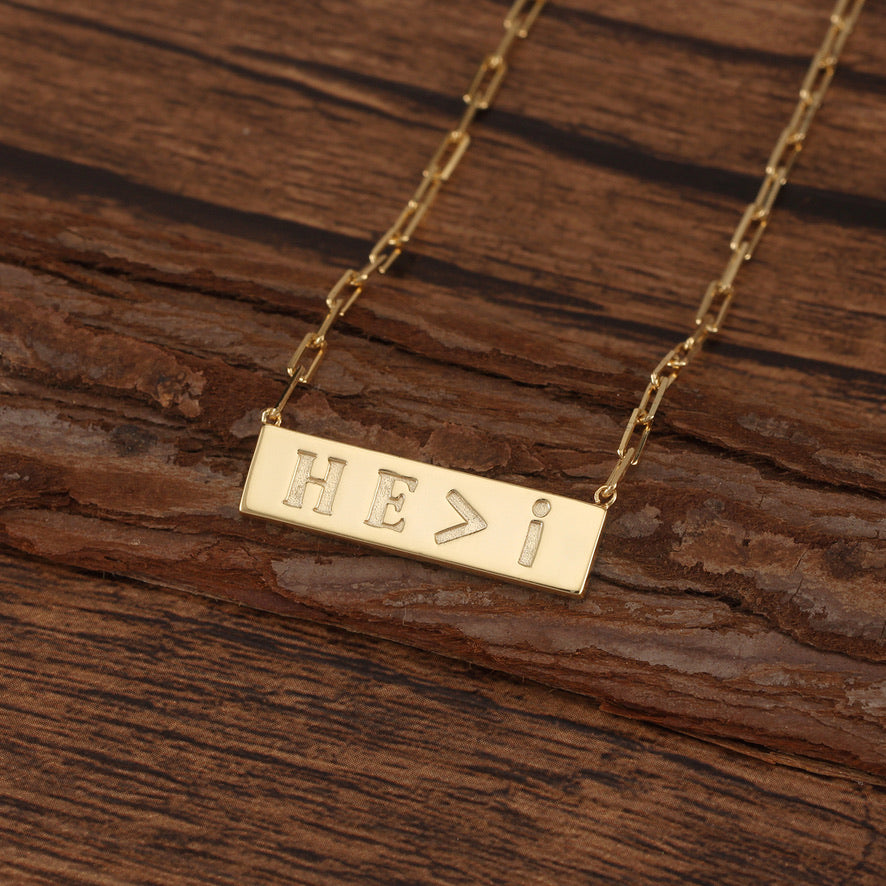 Gold necklace with 'HE>I' Engraved on a bar pendant on a wooden surface