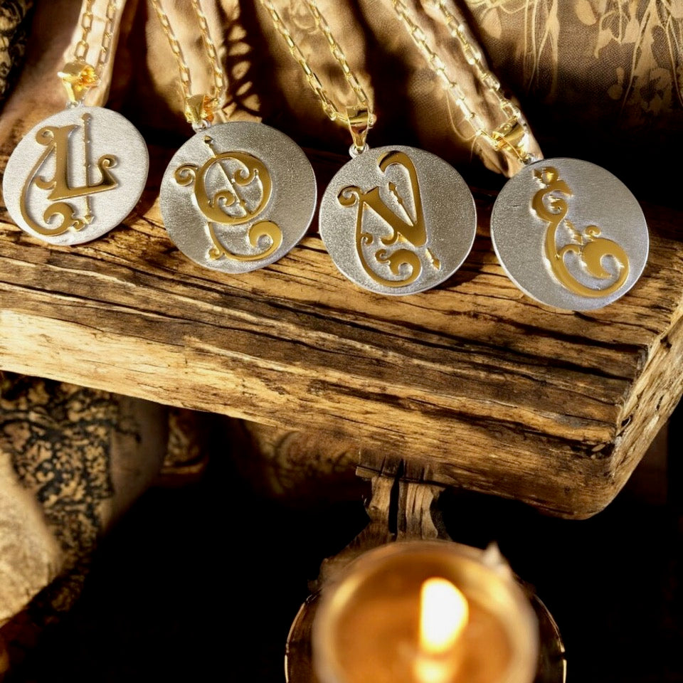 Saints & Saviors Fancy Initial Pendant Necklace A-Z, gold and silver two-tone necklaces with initials L, O, V, E, displayed on a wooden background