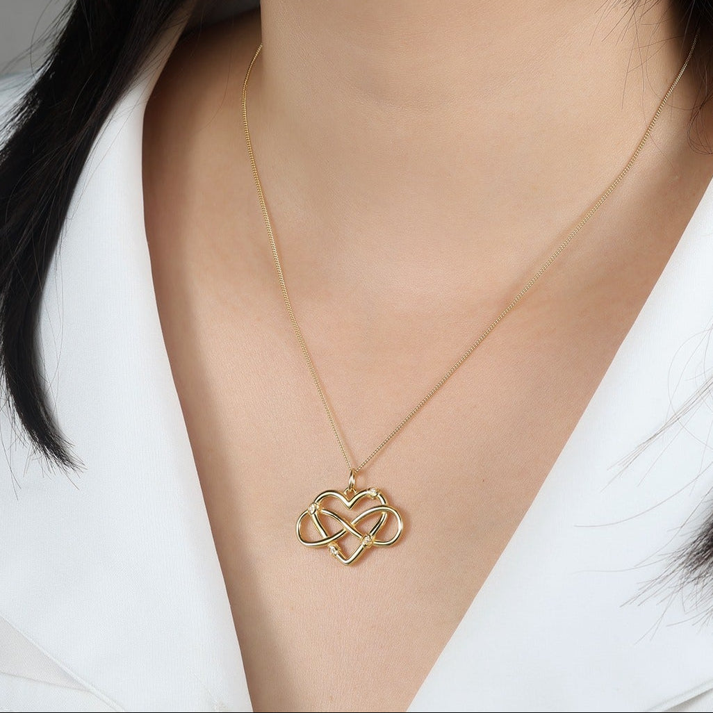 Woman wearing a gold necklace with a heart-shaped and infinity pendant against a neutral background