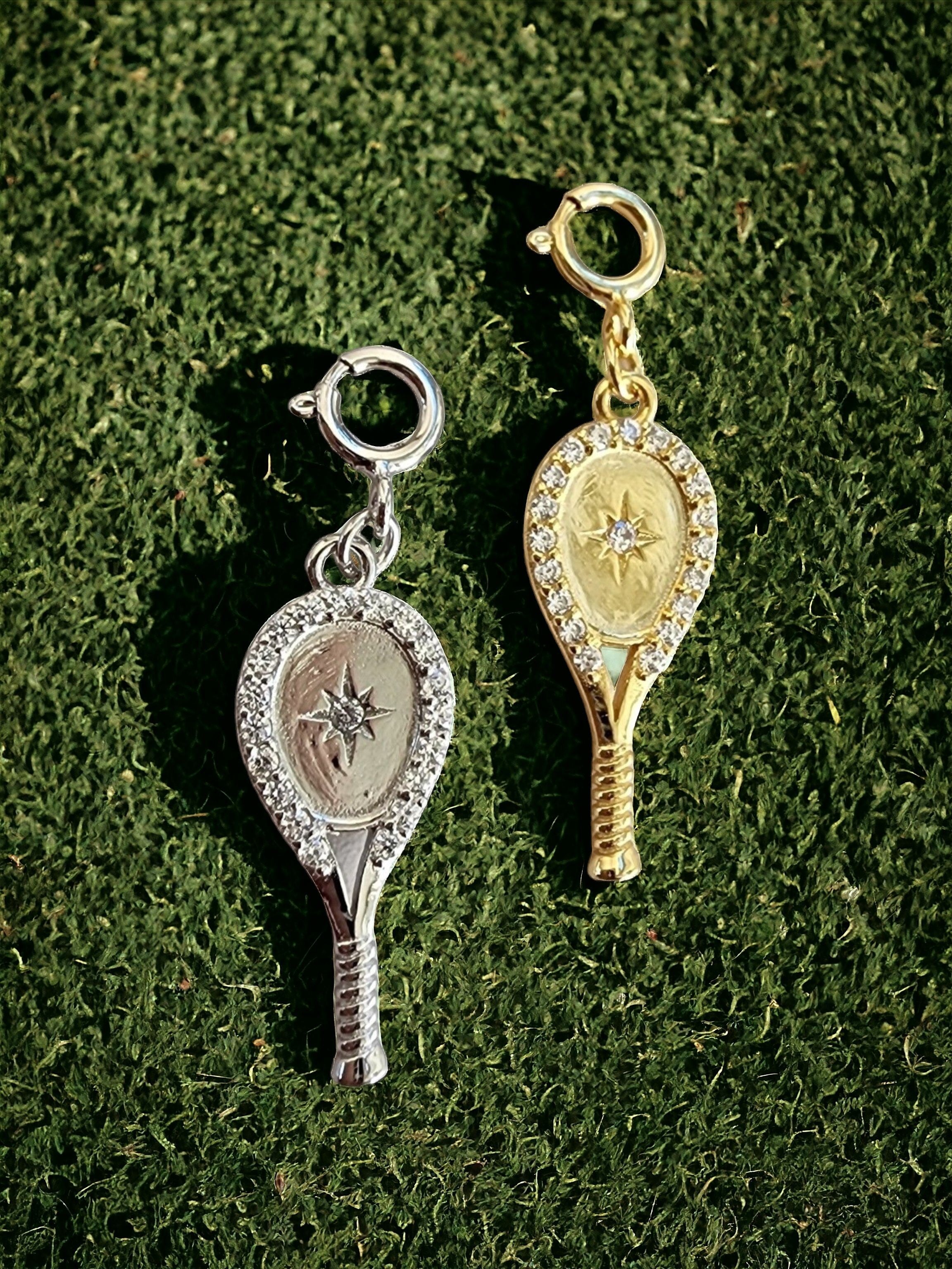 Two tennis racquet-shaped clip-on charms, one silver and one gold with crystals, on a green textured background.