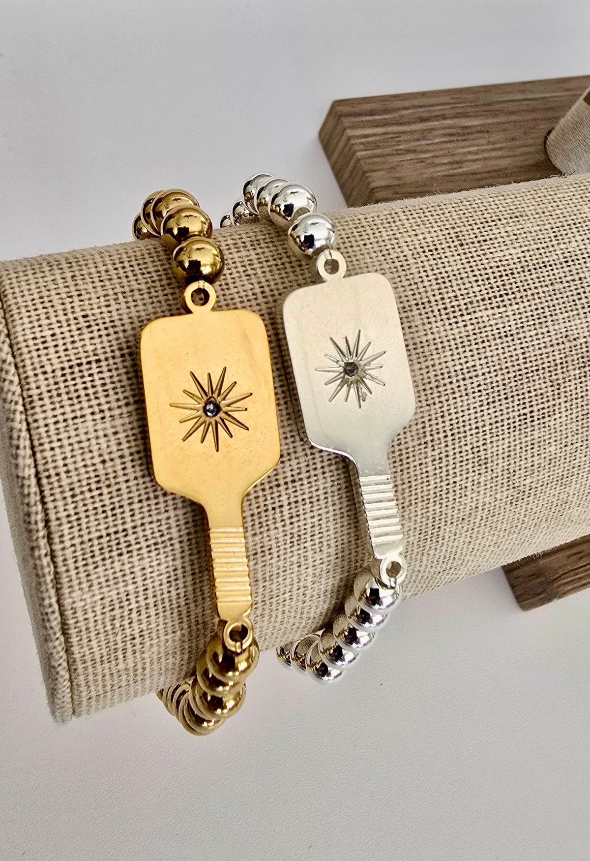Two Picklebelle beaded stretch bracelets with pickleball paddle charms on linen display one each silver and gold