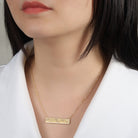 Gold necklace with a bar pendant worn by a person against a neutral background