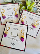 Ruby July Birthstone earrings with gold accents on branded cards, displayed on a light surface.