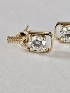 A close-up of a gold pickleball paddle stud earring with a clear round-cut gemstone in a bezel setting, shown on a light gray textured background with another earring partially visible in the background.